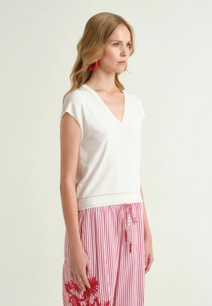 Donna che indossa un top bianco senza maniche con scollo a V e pantaloni a righe rosse e bianche con ricamo floreale, in piedi davanti a uno sfondo chiaro e uniforme.