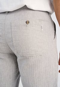 Ljusgrå herringbone-mönstrade byxor med en bakficka som har en metallknapp. Texturerad tyg med en subtil, vävd look.