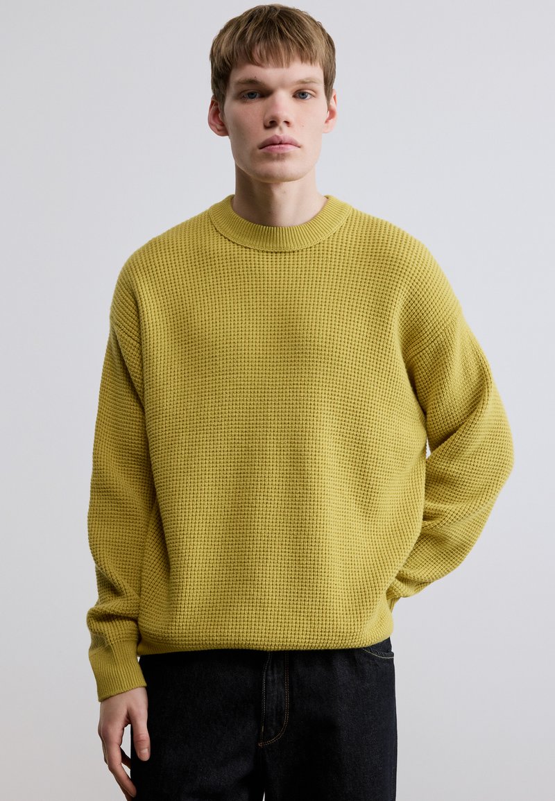 Pull jaune tricoté avec un motif texturé, col rond et coupe décontractée. Associé à un jean en denim foncé.
