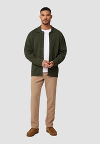 Olijfgroene cardigan met een kraag, knoopsluiting en textielstof. Gedragen over een witte T-shirt en gecombineerd met een beige broek en bruine schoenen.