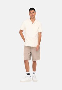 Camisa polo crema de manga corta, con un bolsillo en el pecho, combinada con pantalones cortos de color marrón claro. Zapatillas blancas con calcetines de rayas negras.