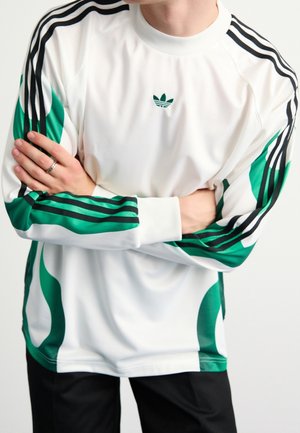 Person iført en hvid adidas langærmet trøje med grønne og sorte striber, armene krydsede, stående mod en ensfarvet baggrund.