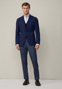 Man i marinblå blazer, ljusblå skjorta, svart väst, grå rutig byxor och mörkbruna dressade skor som står mot en enfärgad bakgrund.