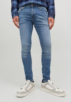 Homme portant un jean skinny bleu clair, des baskets blanches et grises, et une chemise à carreaux bleue, les mains détendues le long du corps.