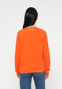 Felpa arancione con scollo rotondo e maniche lunghe, con un logo sottile in bianco sulla schiena. Abbinata a jeans blu.