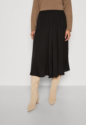 Femme portant une jupe midi noire plissée, un pull beige et des bottes beiges à talons montant jusqu'aux genoux, debout devant un fond uni.