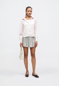 Blusa cor-de-rosa em cetim com gola apontada, combinada com uma saia cinza plissada. Modelo segura uma pequena bolsa branca e usa mocassins escuros.