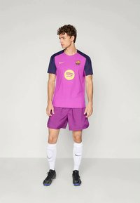 Nogometni dres s kratkim rukavima u svijetlopink bojama s tamnoplavim rukavima i žutim Spotify logotipom, uparen s ljubičastim hlačicama.