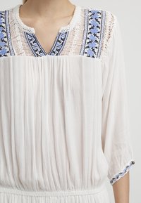 Vit blus med texturerad tyg, med en broderad ok i blått och svart, spetsdetaljer och trekvartsärmar med elastisk fåll.