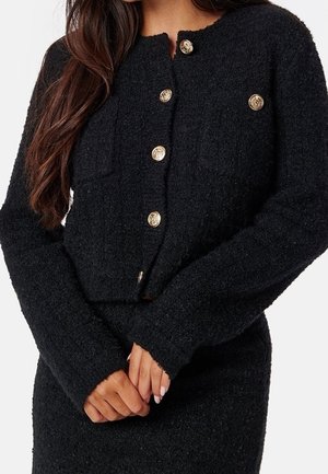 Frau trägt eine schwarze, strukturierte Langarmstrickjacke mit goldenen Knöpfen und passendem Rock, die Hände vor dem Körper verschränkt.