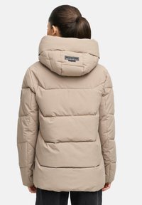 Vrouw met donker haar in een staart, gekleed in een beige gewatteerde pufferjacket met capuchon, gezien van achteren tegen een effen achtergrond.