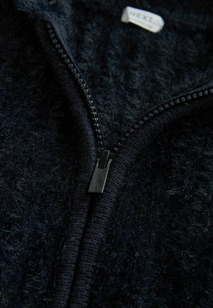 Musta vetoketjullinen vaate, jonka pintaa koristaa pörröinen tekstuuri ja joka on varustettu erottuvalla sileällä vetoketjulla. Kauluksen lähellä on 'NEXT'-merkki. Lähikuva.