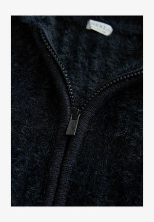 Geacă neagră cu fermoar, cu o textură pufoasă și fermoar neted în contrast. Are o etichetă 'NEXT' aproape de guler. Vedere de aproape.