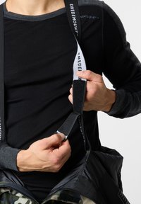 Musta pitkähiha shirt harmailla yksityiskohdilla. Yksilö säätää tarranauhalevyä camo-kuosisessa takissa.