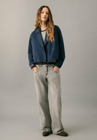 Chaqueta azul marino recortada de doble botonadura sobre un suéter gris, combinada con jeans grises de pierna ancha y calzado marrón, con un fondo neutro.