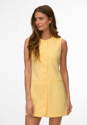 Kobieta o długich brązowych włosach, ubrana w żółtą, bezrękawną sukienkę na guziki, stoi na jednolitym białym tle, patrząc w bok.