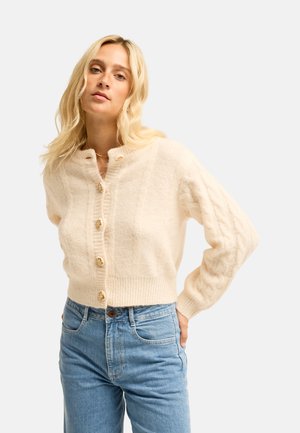 Femme blonde portant un cardigan crème en maille torsadée boutonné et un jean bleu clair taille haute, debout avec les mains derrière le dos.