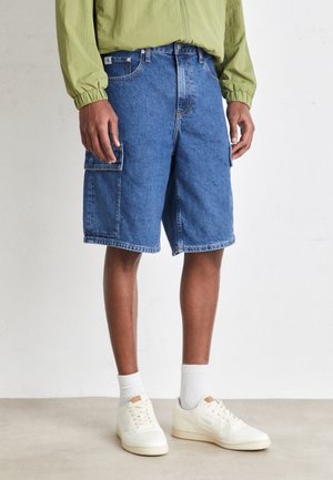 Pantaloni cargo blu in denim con tasche laterali e vestibilità ampia, abbinati a sneakers bianche e calze grigie chiare. Tessuto testurizzato con un design classico.