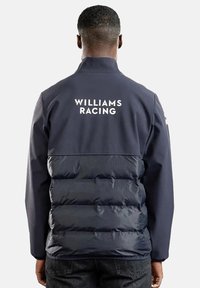 Granatowa kurtka puchowa z gładką górną częścią i pikowanym dolnym fragmentem. Posiada białe logo "WILLIAMS RACING" na plecach. Zamek błyskawiczny na całej długości.