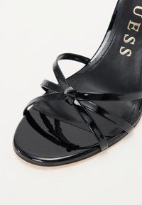 Sandal de tacón alto de charol negro con diseño de tiras cruzadas, que presenta una base de dedo suave y un acabado brillante. Detalle del logo en el interior.
