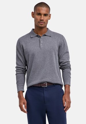 Mann trägt ein graues, langärmliges Poloshirt, das in marineblaue Hosen mit braunem Gürtel gesteckt ist, steht vor einem einfarbigen Hintergrund.