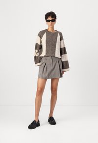 Maglione lavorato a maglia con motivo a treccia in marrone e crema, abbinato a una mini gonna a pieghe a scacchi, indossato con mocassini neri e occhiali da sole.