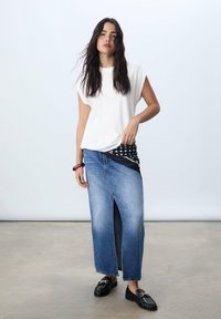 Top bianco a maniche corte abbinato a una lunga gonna di jeans con spacco frontale. Gli accessori includono un foulard bianco e nero a pois e mocassini neri.