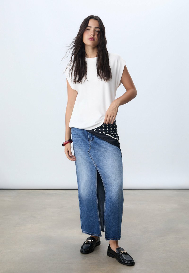 Top bianco a maniche corte abbinato a una lunga gonna di jeans con spacco frontale. Gli accessori includono un foulard bianco e nero a pois e mocassini neri.