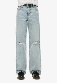 Jean déchiré bleu clair avec une coupe loose, présentant des détails effilochés aux genoux et une coupe droite. Associé à des chaussures noires et une ceinture noire.