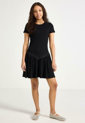 Nuori nainen kiharat hiukset, päällään musta lyhythihainen mekko, jossa on liehukehelma, ja valkoiset kuvioidut tasapohjaiset kengät, seisomassa valkoista taustaa vasten.