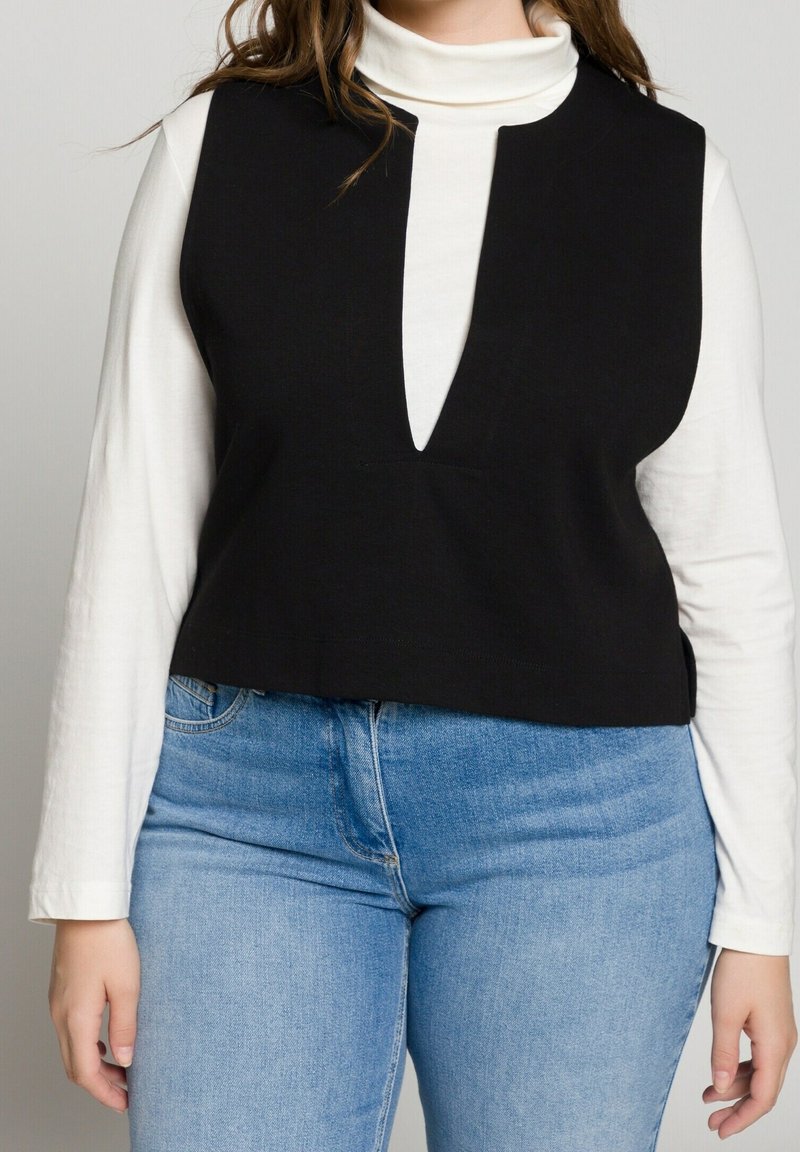 Svart ärmlös topp med V-formad halsringning, bar över en vit långärmad skjorta, kombinerad med blå denimjeans. Slät tygtextur.