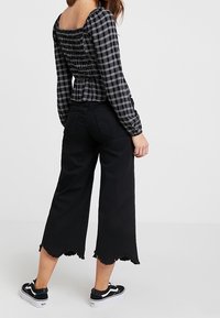 Blusa nera a quadri con schiena arricciata, abbinata a pantaloni larghi neri a culotte con orlo sfilacciato, indossati con sneakers nere.