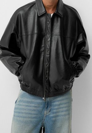 Person iført en sort læderjakke med lynlås med hænderne i lommerne, kombineret med lyseblå denimjeans, mod en grå baggrund.