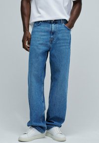 Vaqueros de mezclilla azul claro con un corte holgado, diseño de cinco bolsillos y detalles de costura visibles, combinados con zapatillas blancas.