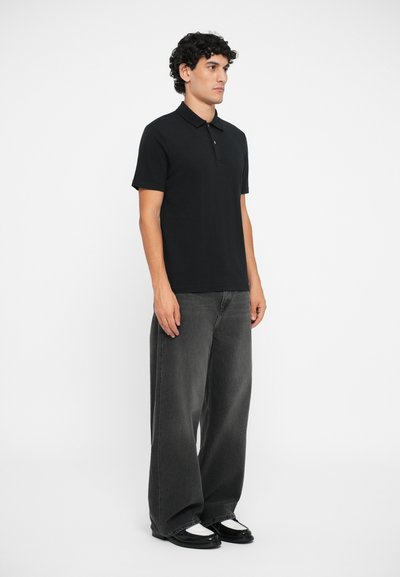 Mustade lühikeste varrukatega polo särk, millele on sobitatud laiade säärehargitega hallid teksad ja mustad kingad valgete aktsentidega. Minimalistlik disain, sile tekstuur.