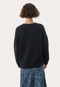Maglione nero oversize in maglia, con scollatura rotonda, orlo a coste e tessuto testurizzato, indossato sopra una gonna blu con motivo.