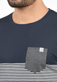 Camiseta de color azul marino con una sección inferior a rayas y un bolsillo sólido. El bolsillo presenta una tela gris texturizada con una etiqueta de logo.