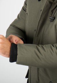 Doudoune olive verte avec une texture lisse, dotée de poignets côtelés noirs et d'une fermeture boutonnée. Comprend un cordon de serrage ajustable au niveau de la capuche.