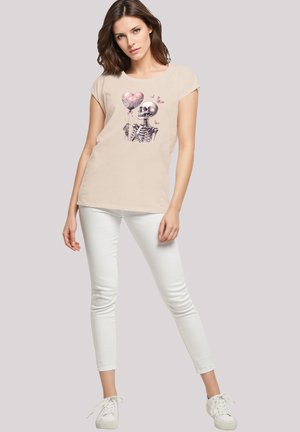 Beige T-Shirt mit kurzen Ärmeln und einem Skelett-Graphic, das einen herzförmigen Ballon hält, kombiniert mit weißen Skinny-Jeans und weißen Sneakers.