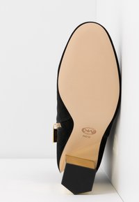 Botín de ante negro con tacón grueso acentuado en dorado, suela beige lisa y detalle de cremallera lateral. La forma hexagonal del tacón añade estabilidad.