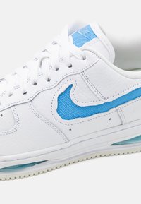 White leather sneaker with a textured blue mesh swoosh design, featuring white laces, a cushioned air sole, and a rubber outsole pattern.