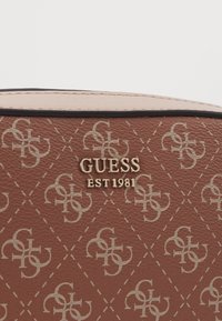 Brązowa syntetyczna torebka z diamentowym wzorem i jasnobeżowymi logo, złotym napisem „GUESS EST 1981” oraz kontrastującymi skórzanymi akcentami.