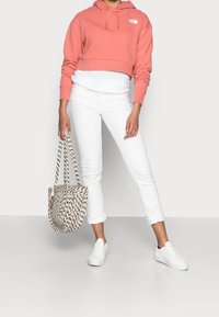 Koraalgekleurde cropped hoodie, witte lange mouwen shirt eronder, witte opgerolde jeans, witte sneakers en een kleurrijke draagtas.