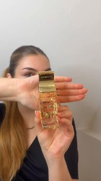 Femme tenant un flacon de parfum transparent avec un bouchon doré, couvrant partiellement son visage avec la main sur un fond uni.