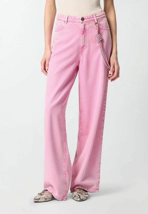 Donna che indossa jeans rosa a vita alta e gambe larghe con una cintura con nodo in corda e scarpe basse con motivo di pelle di serpente.