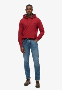 Felpa rossa con cappuccio e tasca a marsupio, indossata sopra una maglietta blu. Abbinata a jeans azzurri e stivali marroni.