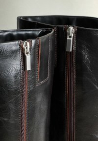 A fekete bőrcipők két oldalsó cipzárral rendelkeznek, ezüst hardware-rel és barna varrással. A felület sima, enyhén gyűrött megjelenésű.