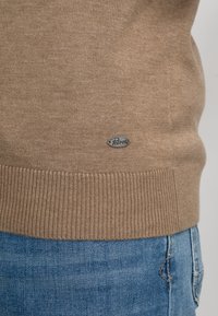 Bruine gebreide trui met een geribde zoom, voorzien van een klein zilveren logopatch. Gedragen over blauwe denim jeans met een gestructureerde afwerking.