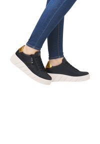 Baskets noires avec des accents dorés, tige texturée, détail zippé et semelle blanche épaisse. Portées avec un jean slim-fit bleu.