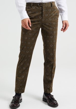 Mężczyzna ubrany w brązowe, wzorzyste, dopasowane spodnie, czarne skarpetki, ciemnobrązowe mokasyny z frędzlami oraz białą koszulę z długim rękawem.